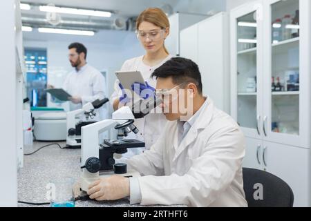 Asian young man working in laboratory together with colleagues. Sits at a desk, studies and examines materials and samples under a microscope. Stock Photo