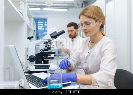 A young woman works in a laboratory, sits at a table with equipment and works on a laptop. Behind, a male colleague is using a tablet. Stock Photo