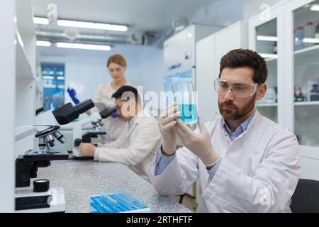 A young male scientist and chemist sits in a laboratory at a table and concentrates on studying a blue substance in a flask. Behind, male and female colleagues are working under a microscope. Stock Photo