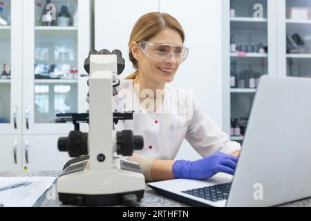 A smiling young woman is working in a laboratory at a laptop. Dressed in a white coat and protective glasses, she sits at her desk. Stock Photo