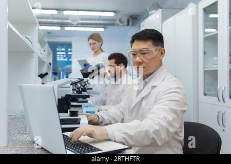 A young Asian man is concentrating on working on a laptop while sitting at a table in a research laboratory. A team of colleagues, a man and a woman working behind a microscope. Stock Photo