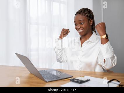 Happy excited african lady entrepreneur sitting at workplace, celebrating success Stock Photo