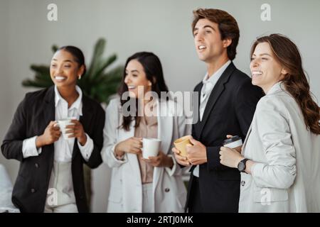 Group of happy international colleagues having break, drinking coffee, holding cups and talking in office interior Stock Photo