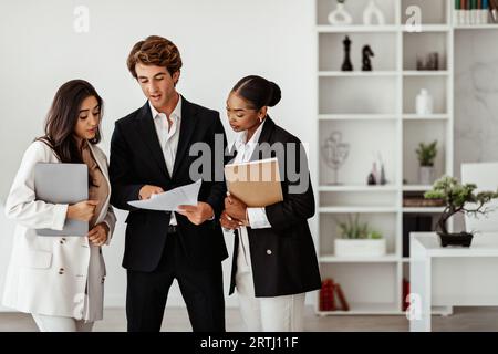 Three multiracial business colleagues reading papers and discussing new project standing in office interior, free space Stock Photo
