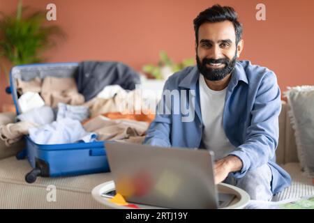 Happy Indian Young Man Using Laptop Near Unpacked Suitcase Indoor Stock Photo