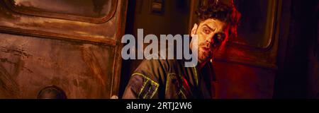 frightened man looking away near rusty carriage in dark underground tunnel with red light, banner Stock Photo