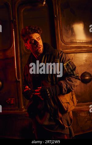 worried man feeling danger and looking away near rusty carriage in post-disaster subway in red light Stock Photo