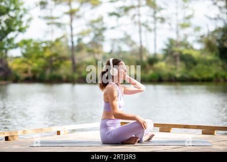 Sporty woman sitting on a jetty by a lake, resting after exercise listening to music on headphones, part of series. Stock Photo