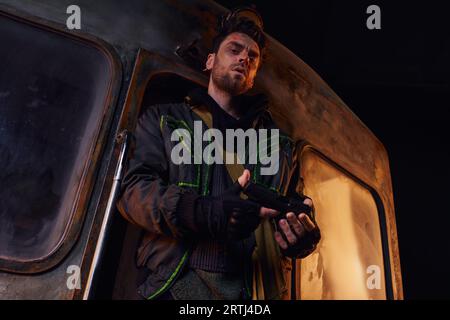 low angle view of unshaven man in worn jacket looking at gun near rusty carriage in abandoned subway Stock Photo