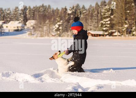 Child playing tug-of-war game with pet dog pulls flying disc in deep snow on winter day Stock Photo