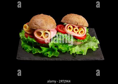 Round baguette sandwich features cheese, ham, tomatoes, and salad on black background Stock Photo