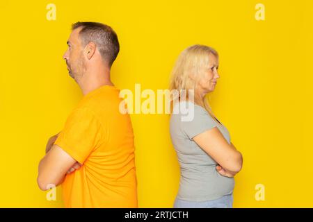 Portrait of a real Caucasian couple in their 40s having an argument, they are angry and turn their backs on each other. Isolated on yellow background. Stock Photo