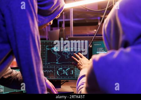 Hackers spying online and checking confidential data on computer screen. Two criminals in hoods collaborating, attacking on server password and gathering sensitive information Stock Photo