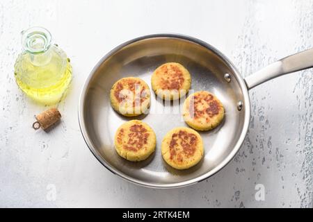 Vegan tofu syrniki with rice flour in a frying pan on a light blue background, top view. Cooking stage. Stock Photo