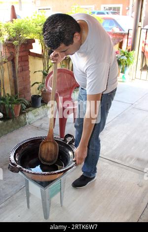 Latino adult man prepares Mexican mole in a clay pot on the stove in the patio of his house, mixes with a wooden spoon part of the Mexican tradition Stock Photo