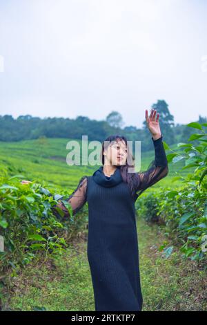 an Asian woman in a black dress is posing in front of a very beautiful tea plantation in the morning Stock Photo
