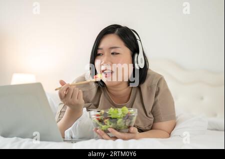 A happy and diet Asian plus-size woman is lying on bed, enjoying her healthy salad while watching a movie on her laptop in her bedroom. Curvy girl dom Stock Photo