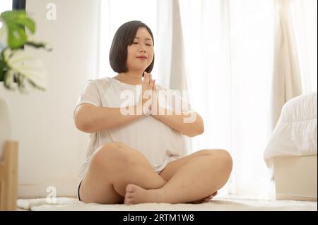 A peaceful and happy Asian curvy plus-size woman in sportswear practicing yoga at home, eyes closed, meditating in her bedroom. Stock Photo