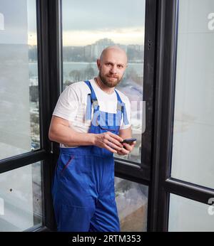 Portrait of young bald male construction worker in blue overalls using smartphone. Man standing near big window in apartment under renovation. Stock Photo
