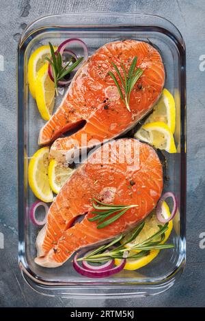 Two steaks of fresh raw red fish of the salmon family, trout pieces with various ingredients in a transparent glass baking dish on the table, top view Stock Photo