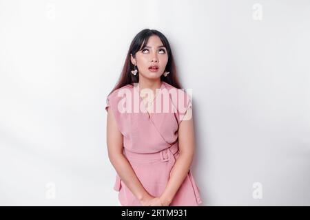 Dissatisfied young Asian woman dressed in pink blouse rolling her eyes and raising hand discontentedly, standing against white background Stock Photo