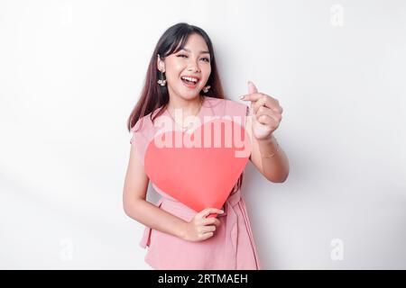 A happy young Asian woman wearing a pink blouse feels romantic shapes heart gesture expressing tender feelings and holding a red heart-shaped paper Stock Photo