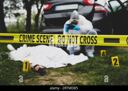 Criminologist taking photo of dead body at crime scene outdoors, focus on yellow tape Stock Photo