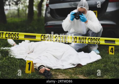 Criminologist taking photo of dead body at crime scene outdoors, focus on yellow tape Stock Photo