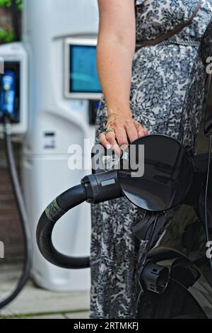 Female hand hold the charging plug and charge electric car at a ev charging point at gas station. Charging screen in the background. Eco friendly Stock Photo