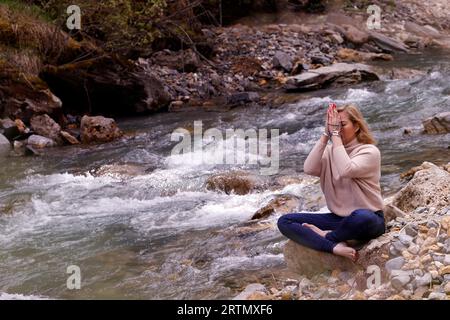 Praying  woman sitting on a rock at the edge of a river. Concept of religion in nature. Stock Photo