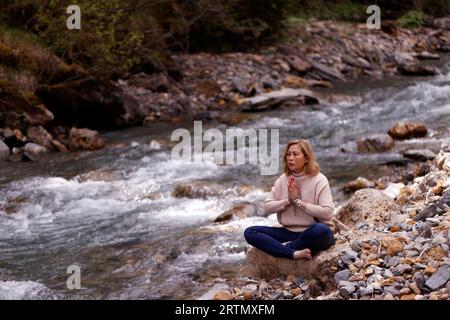 Praying  woman sitting on a rock at the edge of a river. Concept of religion in nature. Stock Photo