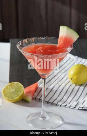 Watermelon margarita served in a martini glass. Lime and watermelon slices rest on a black and white striped cloth sitting on a white marmbled surface Stock Photo