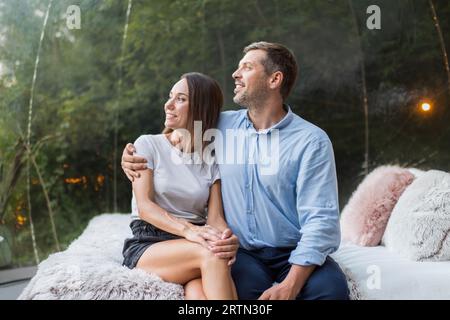 A hugging couple inside transparent bubble tent at glamping while sitting on the bed. Lush forest on the background Stock Photo