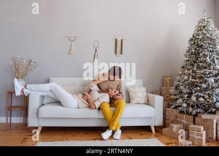 Loving, cheerful couple on a sofa near a Christmas tree with gifts sits on a white sofa. Crazy lovers spend the holidays together. Stylish, trendy int Stock Photo