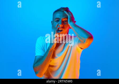 Portrait of young man, student on blue studio background in neon light, tired covering face hands, depression and sadness, upset and irritated for Stock Photo