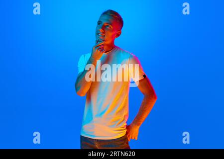 Portrait of young man, student on blue studio background in neon light, filter. Handsome, dreaming male model in casual style. Stock Photo