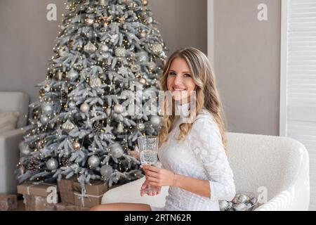 Portrait of trendy, stylish young woman, millennial, with glass of champagne in hands near Christmas tree, sitting on white chair. Girl is happy, smil Stock Photo