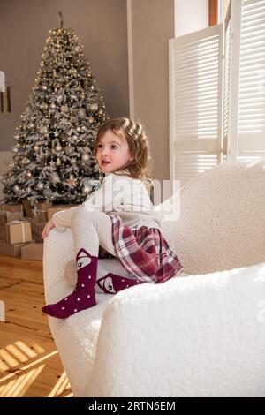 Funny little girl lies in a soft white chair, fooling around, making faces near the Christmas tree. Child in a cozy atmosphere at home. Festive interi Stock Photo