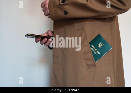 Translation: 'military id'. Civil man holds smartphone in one hand and has ukrainian military id in coat pocket. Mobilization, law, Ukraine Stock Photo