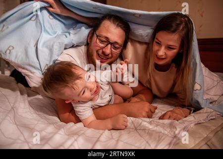 Cheerful, cute family is hiding with baby son under blanket. Mother and father try to catch an active baby, boy funny runs away from parents. Young ma Stock Photo
