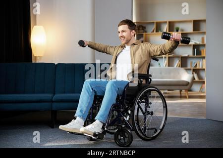 Physical rehabilitation for disabled people. Happy guy in wheelchair making exercises with dumbbell at home. Cheerful impaired young man working out w Stock Photo