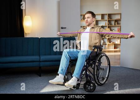 Young disabled man in wheelchair doing exercises with rubber band at home. Caucasian handicapped guy working out in living room. Stock Photo