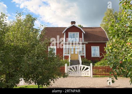 a typical red and white swedish house in smalland. White garden gate, brown fence. green lawn in the garden. Trees in the foreground. Blue sky with sm Stock Photo