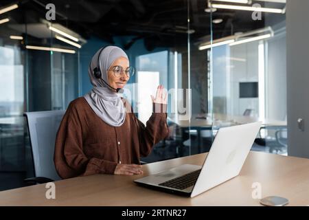 Young Muslim female teacher in hijab and headset doing distance learning online. Sitting at a desk in front of a laptop and smiling and waving to students and pupils. Stock Photo