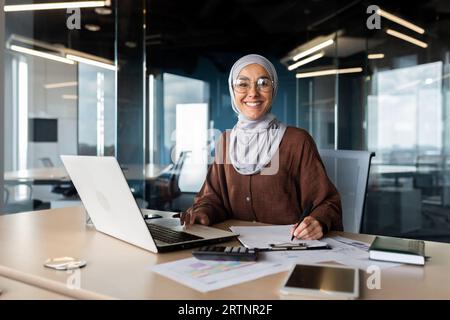 Portrait of young successful Arab woman in hijab working in office with documents and laptop. Smiling and looking at the camera. Stock Photo