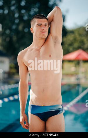A young man wearing a swimsuit stands confidently near a large outdoor swimming pool, looking ahead with a determined expression Stock Photo