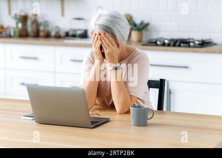 Fatigue concept. Exhausted middle aged caucasian gray haired woman, pensioner, sits at a table with laptop in kitchen, feels overworked and stressed, fatigue from working on a laptop, has migraine Stock Photo