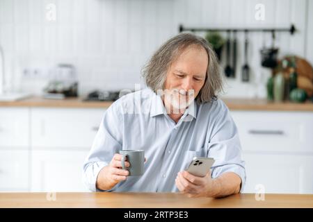Satisfied senior caucasian grey haired man with beard, sits at home in the kitchen at the table, relaxing, drinks coffee, using his smartphone, browsing social media, reading news, watching video Stock Photo