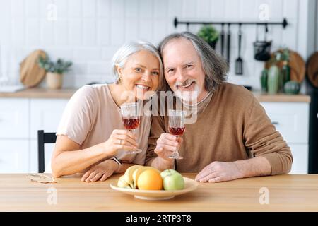 Happy pensioners. Elderly caucasian spouses, spend time together in the kitchen at home, hold glasses of wine in their hands, celebrate an important date, anniversary, look at camera, smile joyfully Stock Photo