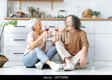 Happy pensioners. Mature caucasian spouses, sits on the floor in the kitchen at home, hold glasses of wine in their hands, celebrate an important date, anniversary, look at each other with love, smile Stock Photo